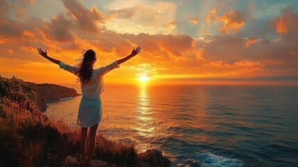 Fototapeta premium Woman standing on cliff edge with arms raised, overlooking vibrant orange sunset over the ocean with dramatic clouds and calm waves