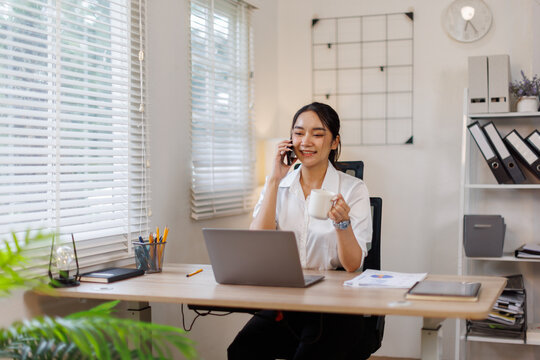 Happy young business woman manager talking consulting client on cell phone working on laptop in the office. Busy woman professional business making call on cell using computer sitting at desk.