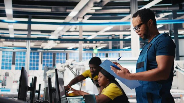 Black engineer in photovoltaics factory reviews performance of solar panels assembly lines, checking paperwork for finetuning. Supervisor inspecting solar cell machinery. Camera B.