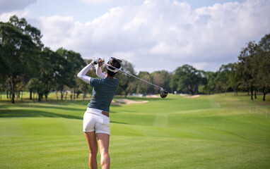 Professional female golfer lush green golf course, mid-swing after hitting a shot through after driving the ball down the fairway trees and distant sand bunkers, typical of a competitive setting.  