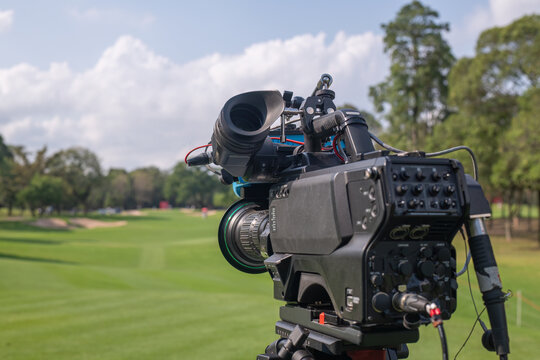 Professional broadcast video camera set up on a tripod overlooking a golf course. The camera is focused on a lush green fairway, likely capturing footage for a live or recorded golf tournament.
