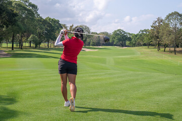 Professional female golfer lush green golf course, mid-swing after hitting a shot through after driving the ball down the fairway trees and distant sand bunkers, typical of a competitive setting.  
