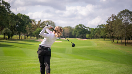 Professional female golfer lush green golf course, mid-swing after hitting a shot through after driving the ball down the fairway trees and distant sand bunkers, typical of a competitive setting.  