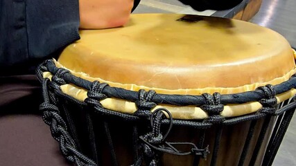 A musician is playing with his bare hands a djembe bongo drum, tapping their hands on the surface to create a rhythm. The camera moves around to capture different angles of their performance.