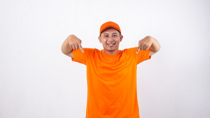 Asian deliveryman in orange t-shirt uniform poses as if he is pointing  something bellow isolated...