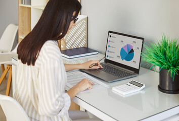 Fototapeta premium Portrait of a concentrated young business woman accountant sitting at the desk and working on a laptop computer at office or at home and looking to the monitor screen. Accounting job and finance