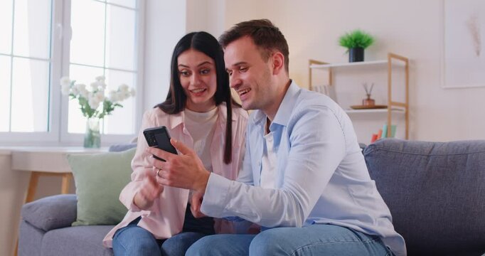 Happy young couple sitting together on sofa in living room, exchanging high five and celebrating success. Smiling man showing something interesting on mobile phone, feeling excited with good news.