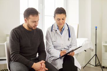 Portrait of a young man patient in medical office listening to a friendly female doctor holding...