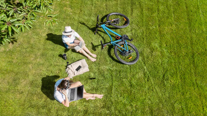 Young girls with bicycle in park, using laptop computer and headphones, two student girls studying and relaxing outdoors sitting on grass near bike, aerial drone view from above
