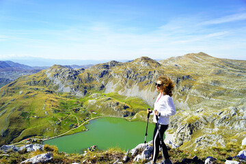 Hiking in the most picturesque places of Montenegro: smiling female tourist with trekking poles walking on the top of the mountain with a magnificent view. Woman with Kapetanovo Lake in the background