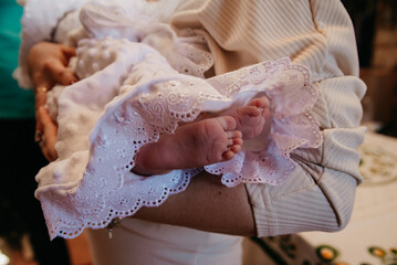 During the rite of baptism of a child in the church, the godmother holds the child dressed in a magnificent dress in her arms, the child's small legs are in the photographer's frame