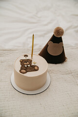 a small cake or cupcake with one candle inserted in it and decorated with a decorative chocolate bear on an isolated background for a birthday celebration and a festive hat on the head next to it