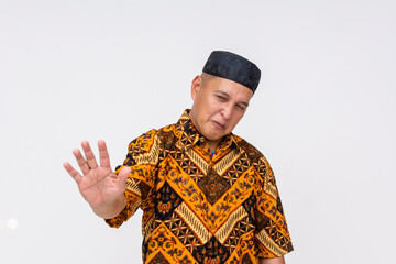 Middle-aged Indonesian man showing discomfort or rejection with a raised hand and squinting expression, wearing Batik shirt and songkok. Isolated on a light background.
