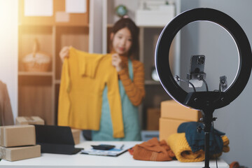 vlogger, blogger small SME business asian beautiful woman using mobile phone video call for sell clothes live stream selling online, show product present detail at home office, entrepreneur ecommerce
