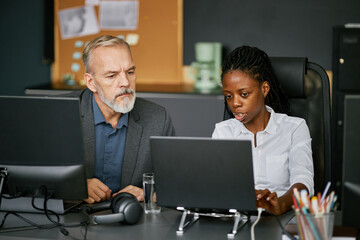 Senior Caucasian man collaborating with young Black woman on computer project in modern office space. Notable technology and gadgets on desk, intense focus on task