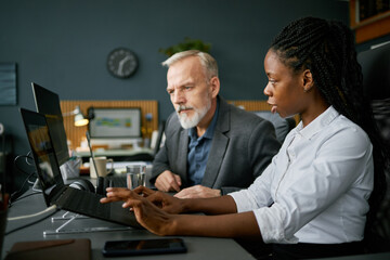 Senior man and younger woman working together on project in modern office setting, using laptops and analyzing data in relaxed yet focused atmosphere