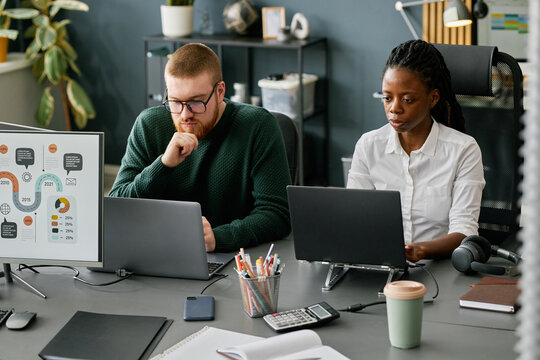 Caucasian man and African American woman engaged in office work, using computers and collaborative tools on desk, discussing business matters