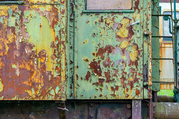 Close-up of a heavily weathered train car door with peeling green and yellow paint, exposing rust and corroded metal underneath. The image captures the texture of decay, aging, and industrial deterior