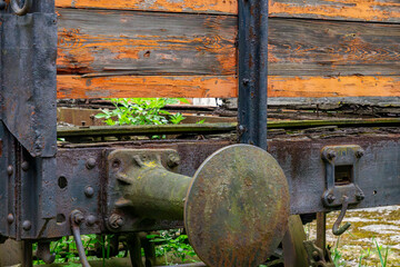 Close-up of rusty elements of an old freight train car, with a weathered wooden body showing cracks and peeling paint. In the background, greenery is growing around the tracks, adding a sense of aband