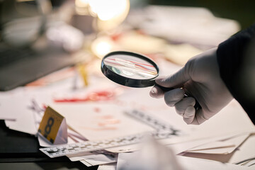 Gloved hand holding magnifying glass examining documents on cluttered workspace filled with papers and laboratory tools, suggesting forensic investigation scene