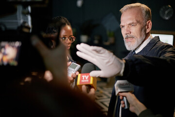 Middle-aged man speaking during press conference while reporters holding microphones. Appears to be...