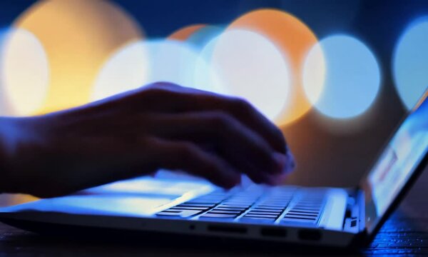 Female hands is typing on a laptop at night. Blurred lights on the background