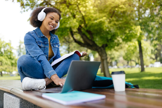 Happy black female student study outdoors with laptop and workbooks, smiling african american woman using computer while sitting on bench in campus, preparing for exams, free space