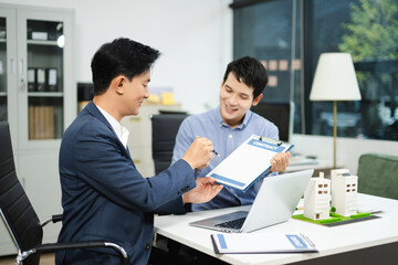 Two businessmen discuss and sign a real estate contract with housing models on a desk for concepts of property, investment