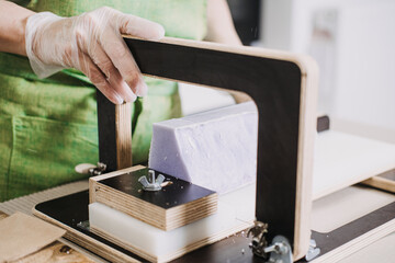 Close-up of a woman slicing handmade purple soap bars using a wire soap cutter. Small-batch skincare, craftpreneur economy, home-based entrepreneurship, creative business models