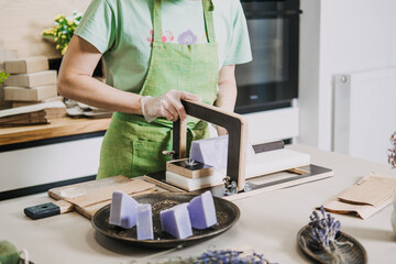 Woman in green apron slicing handmade lavender soap on a worktable with bunches of dried lavender nearby. Clean beauty, natural ingredients, chemical-free soap, small-scale production