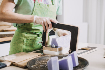 Person in green apron using a wire soap cutter to slice a large block of lavender-colored handmade...