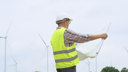 Engineer holding blueprint while surveying wind turbines at renewable energy site under bright sky...