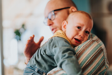 Father holding and soothing a crying baby in a warm, indoor setting, showcasing emotions of care,...