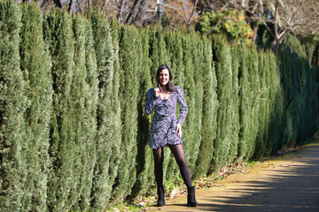 Naklejka premium Portrait of a young, beautiful Latin brunette woman in front of the green hedges separating a road. The woman is happy, making different expressions on the path and having fun.