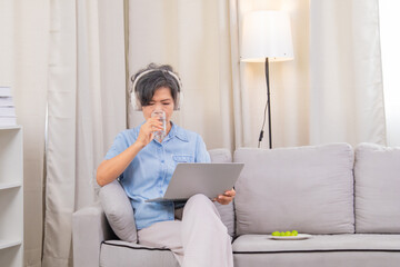 Fototapeta premium Asian senior woman friendly with modern technology lifestyle, cheerful pensioner enjoy using laptop connect to social media listen to music at home, elderly female holiday with computer headphone