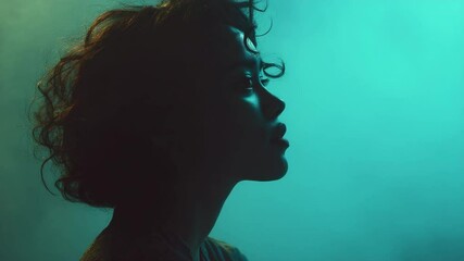Close up profile view of a young woman with dark, curly hair, silhouetted against a teal, smoky background. Moody, dramatic lighting. - Powered by Adobe