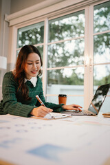A confident Asian businesswoman sits at her desk with a laptop, smiling while analyzing financial...