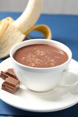 Tasty chocolate milk in cup, pieces and banana on blue table, closeup