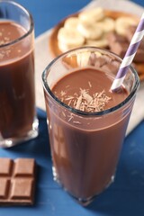 Tasty chocolate milk in glasses, pieces and banana on blue wooden table, closeup
