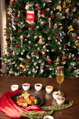 A shot of roasted chicken with vegetables, knife and fork on a brown table with red satin cloth, served with a glass of cocktail, surrounded by Christmas décor and a decorated tree in the background.