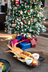 A lobster placed in a plate on a table, with a decorated Christmas tree in the background adorned with stars, bells, and baubles, as warm sunlight filters through the scene.
