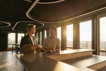 Two men having a business meeting in a modern office during sunset, holding coffee.