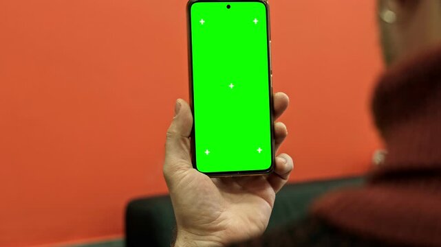 Hand of young man holding smartphone green screen in business office work. Mobile phone vertical mockup. Male person using phone with blank green background display. Chroma key smart phone. Close up.
