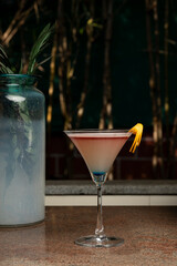 A pink and blue cocktail served in a martini glass with an orange peel garnish, placed on a marble slab with blurred indoor plants and a warm restaurant ambiance in the background. 