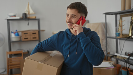 Young man talking on smartphone while holding a cardboard box in a new apartment living room, surrounded by packing items, symbolizing moving process.