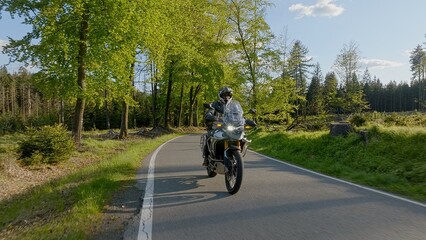 Driver riding motorcycle on empty asphalt road, spring mountains during sunset