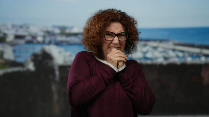 Woman wearing glasses appearing frightened at seaside promenade with ocean backdrop, middle-aged hispanic features, giving a sense of outdoor apprehension and vulnerability.