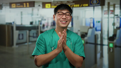 Obraz premium Young chinese man in green medical scrubs clapping at an airport terminal indoors with signs in the background.