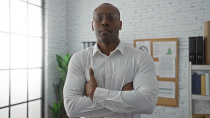Man standing confidently in a bright office setting with arms crossed, wearing glasses and a white shirt, exuding professionalism and focus.