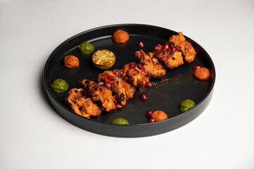 Classic Chicken Tikka marinated in yoghurt and flavorful spices, skewered and cooked in a clay oven. Served with green and red chutney, garnished with anardana, in a black plate on a white background